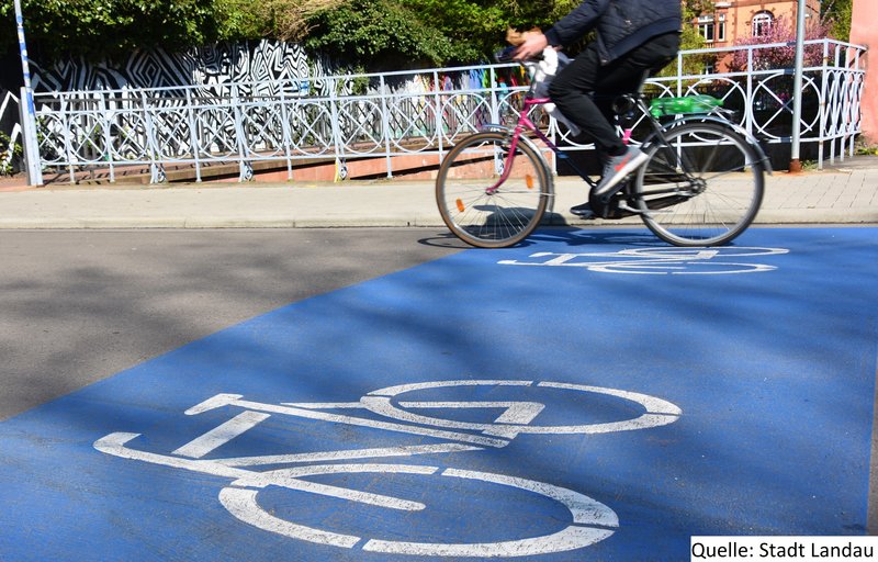 Straße mit Person, die Fahrrad fährt und einer weißen Fahrradmarkierung auf blauem Grund auf dem Boden.