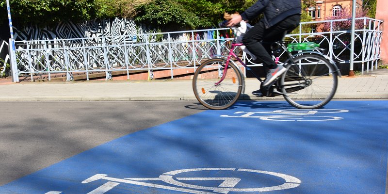 Straße mit Person, die Fahrrad fährt und einer weißen Fahrradmarkierung auf blauem Grund auf dem Boden.