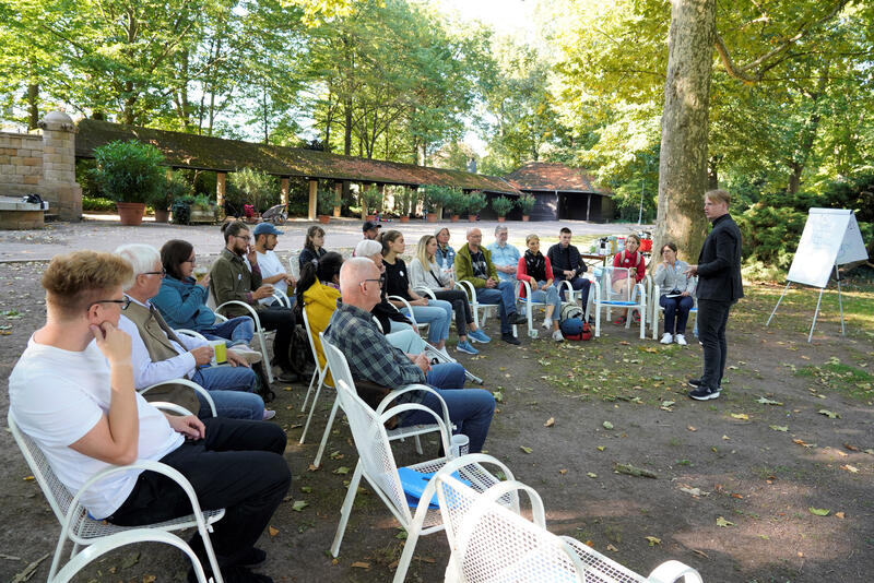 Einige Personen sitzen in einem Park in einem Halbkreis auf Stühlen und hören Bürgermeister Lukas Hartmann zu.